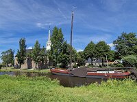 Church and skutsje of the Dutch village Elahuizen, Friesland, Ne  Church and skutsje of the Dutch village Elahuizen, Friesland, Netherlands : church, skutsje, monument, historic, building, architecture, 19th century, religion, religious, tower, landmark, historic freighter, ship, boat, mast, sailing, tree, trees, blue sky, sky, summer, summertime, sun, sunny, outside, outdoor, outdoors, no people, nobody, Friesland, Fryslan, Netherlands, Dutch, europe, Elahuizen, De Friese Meren, sailing ship